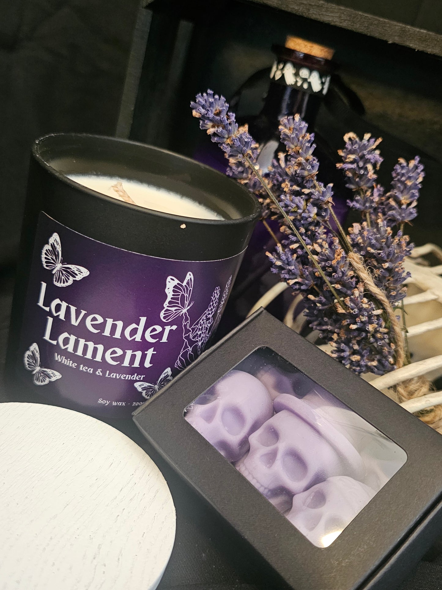 a candle in a black container beside a box of skull shaped wax melts, both on a dark background with lavender buds and a purple bottle