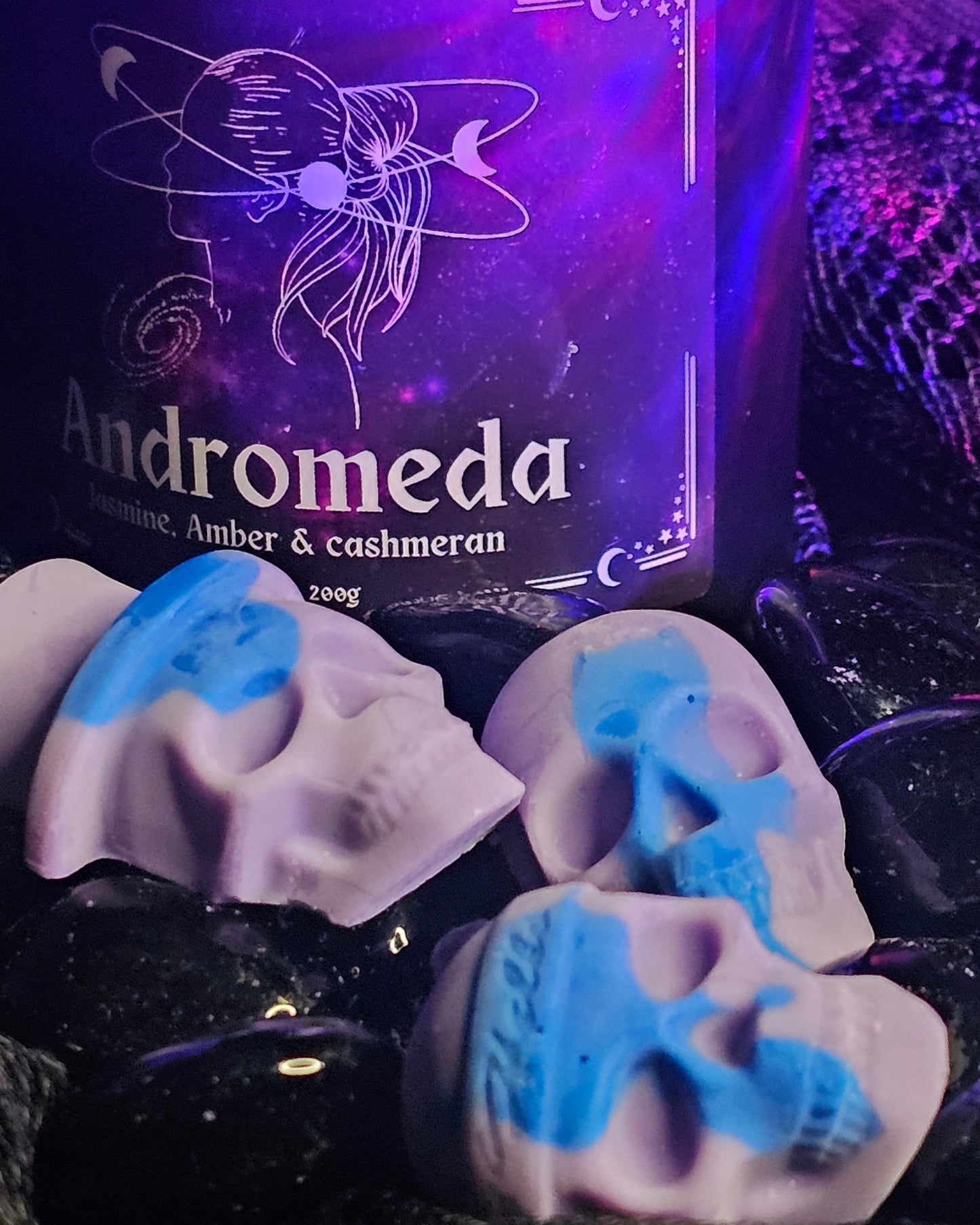 3 skull shaped wax melts laying infront of an Andromeda scented candle, both on a dark rocky background