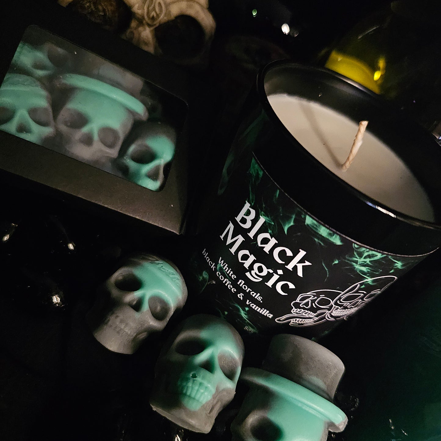 A black candle jar with 'Black Magic' written on it, along with mentions of white florals, black coffee, and vanilla. The background features green and black skull shaped wax melts