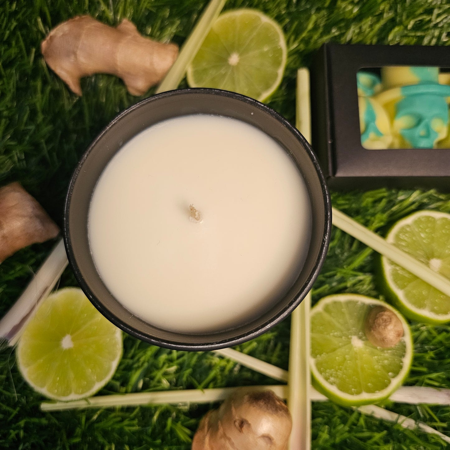 A candle veiwed from above, surrounded by lemongrass leaves, ginger roots, lime slices and yellow and green Skull shaped wax melts lay alogside the candle on a grassy background.
