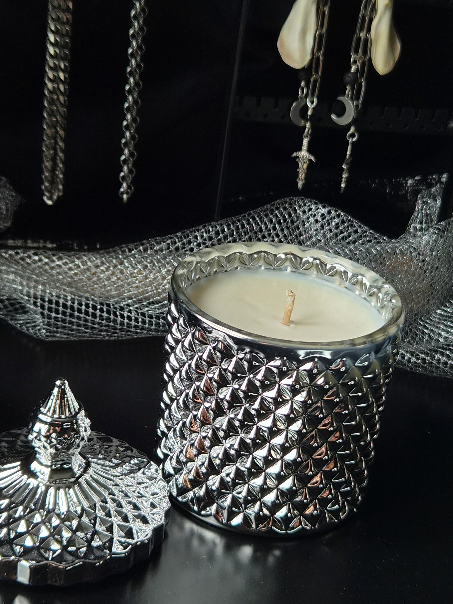 A silver textured candle jar with a matching lid, filled with white wax, displayed on a dark surface with gothic jewelry and mesh in the background.