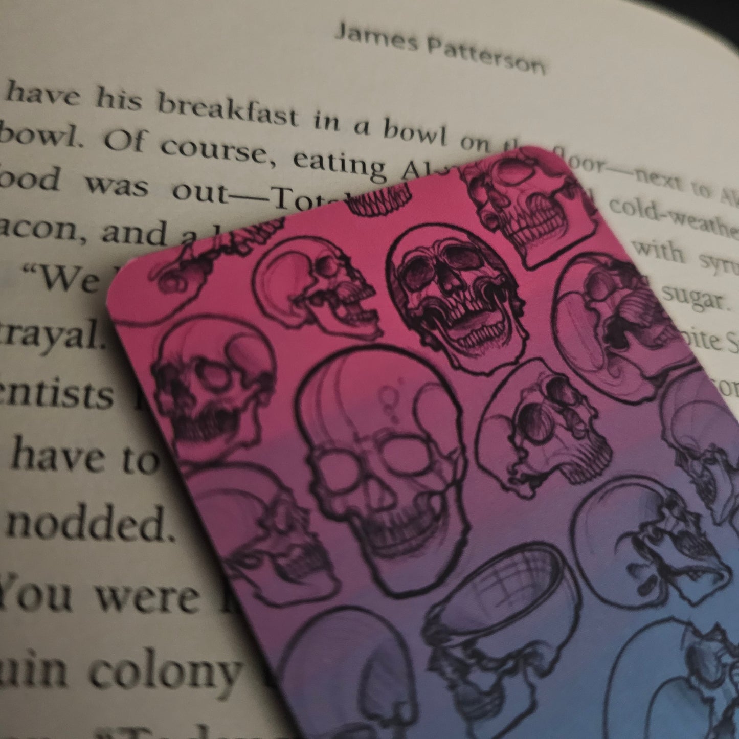 A bookmark with a gradient of pinks and blues, featuring hand-drawn skulls in various forms on it, placed on an open book, close up