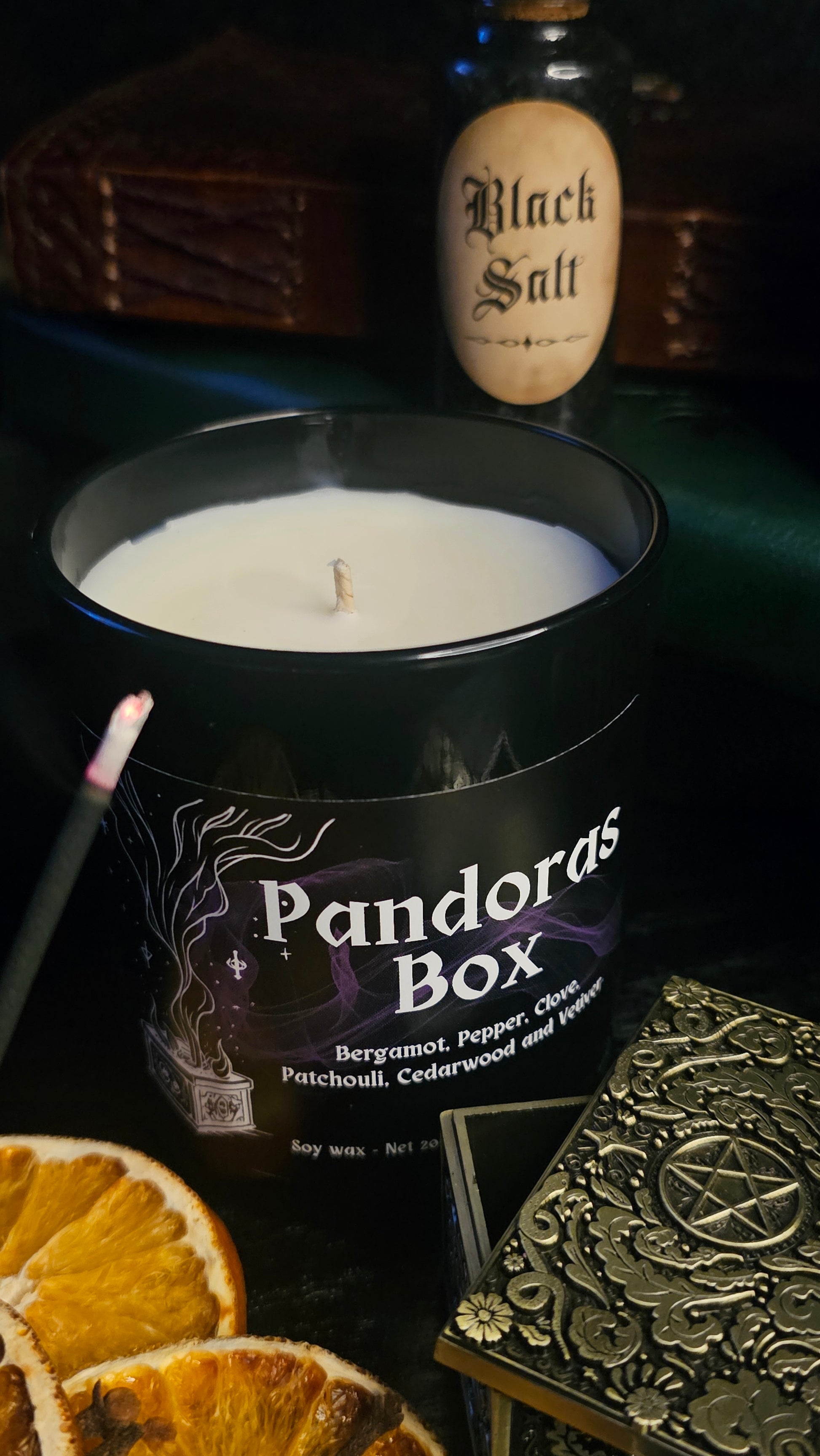 Candle in a 'Pandora's Box' container with a bottle labeled 'Black Salt' and dried oranges on a dark surface.