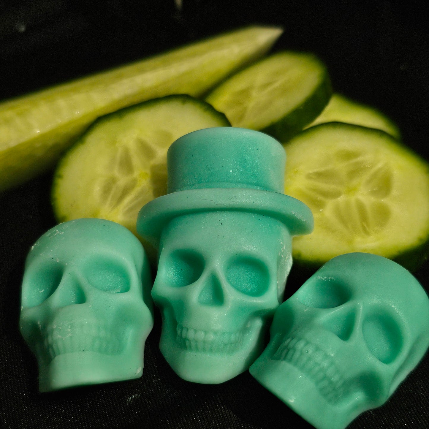 Close up image of 3 green skull shaped wax melts laying on a dark background with cucumber slices 