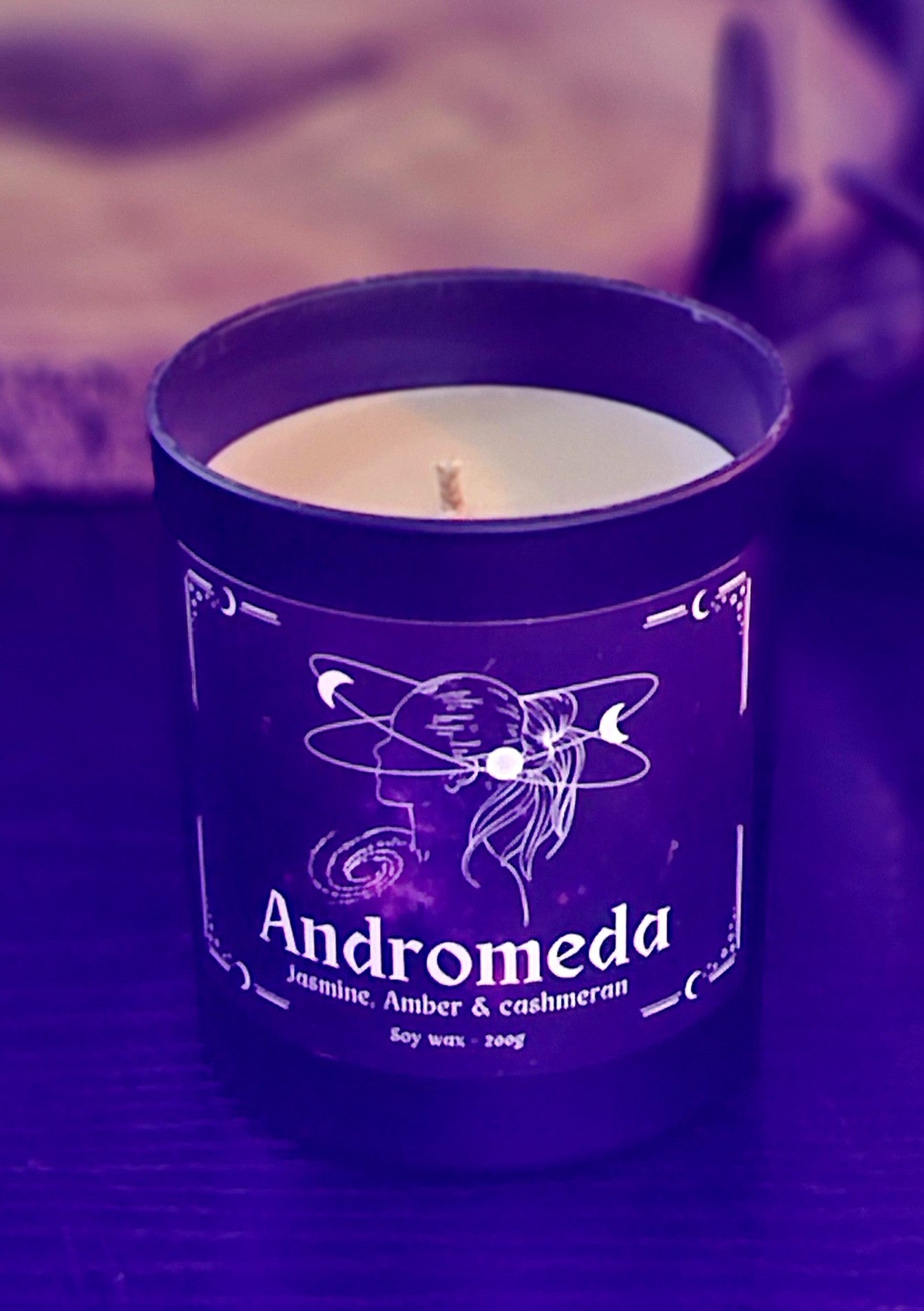Candle in a black container labeled 'Andromeda' with a purple tint and blurred background