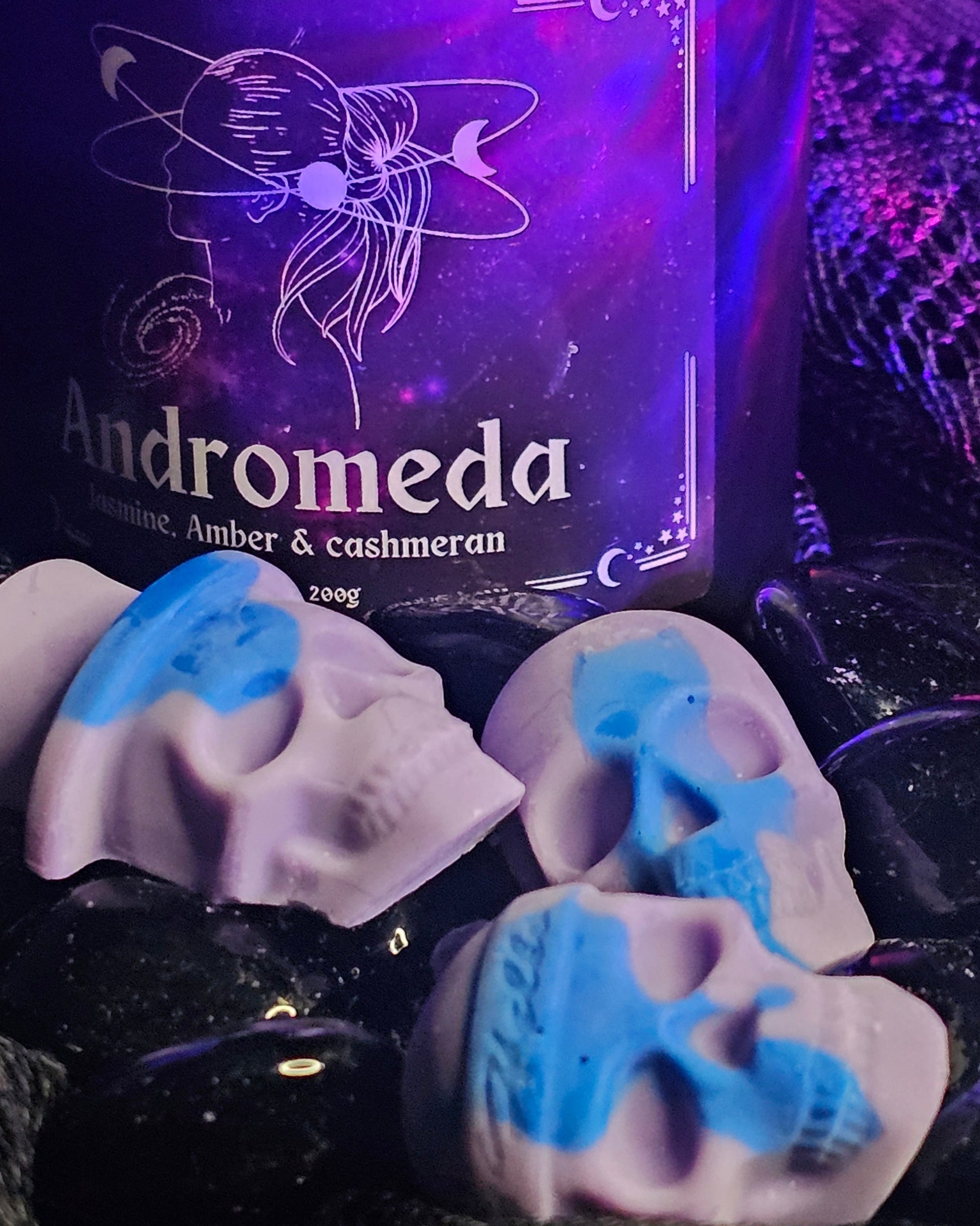 3 skull shaped wax melts laying infront of an Andromeda scented candle, both on a dark rocky background 