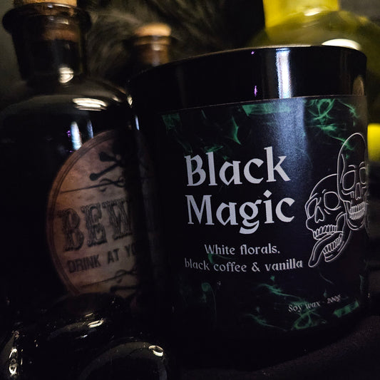 A black candle jar with 'Black Magic' written on it, along with mentions of white florals, black coffee, and vanilla. The background features other dark colored bottles and containers.