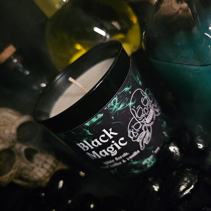 A Candle surrounded by glass bottles and ornimental skulls with smoke rising from the surface it is resting on.  