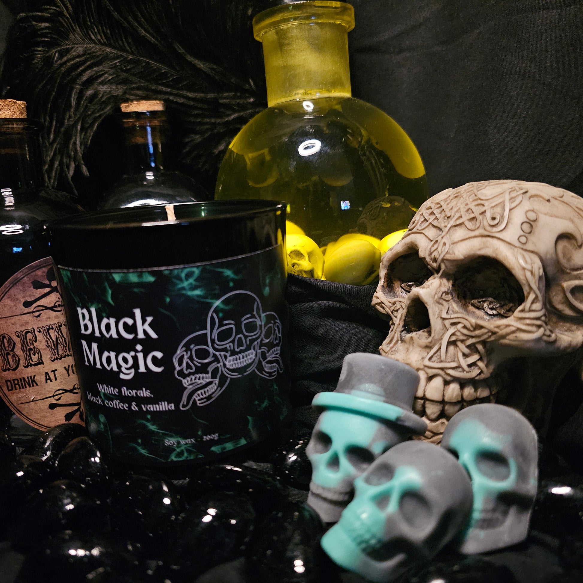 Three Skull shaped wax melts beside a black candles container labelled "Black Magic" On a dark background with various alchemist style bottles 