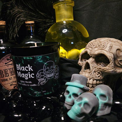 Three Skull shaped wax melts beside a black candles container labelled "Black Magic" On a dark background with various alchemist style bottles 