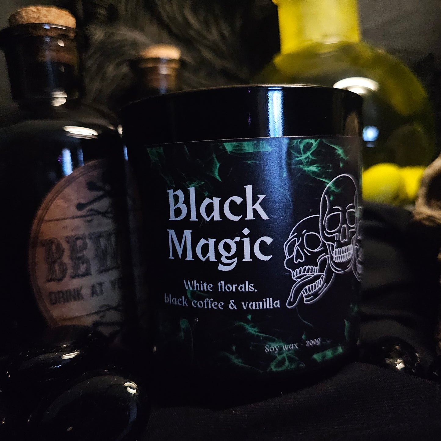A black candle jar with 'Black Magic' written on it, along with mentions of white florals, black coffee, and vanilla. The background features other dark colored bottles and containers.