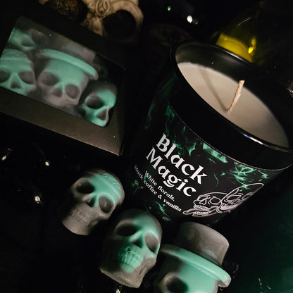 A black candle jar with 'Black Magic' written on it, along with mentions of white florals, black coffee, and vanilla. The background features green and black skull shaped wax melts 