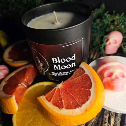 Candle in a black jar with a label the reads "Blood Moon - Blood orange, rosehip, orange blossom and oakwood"