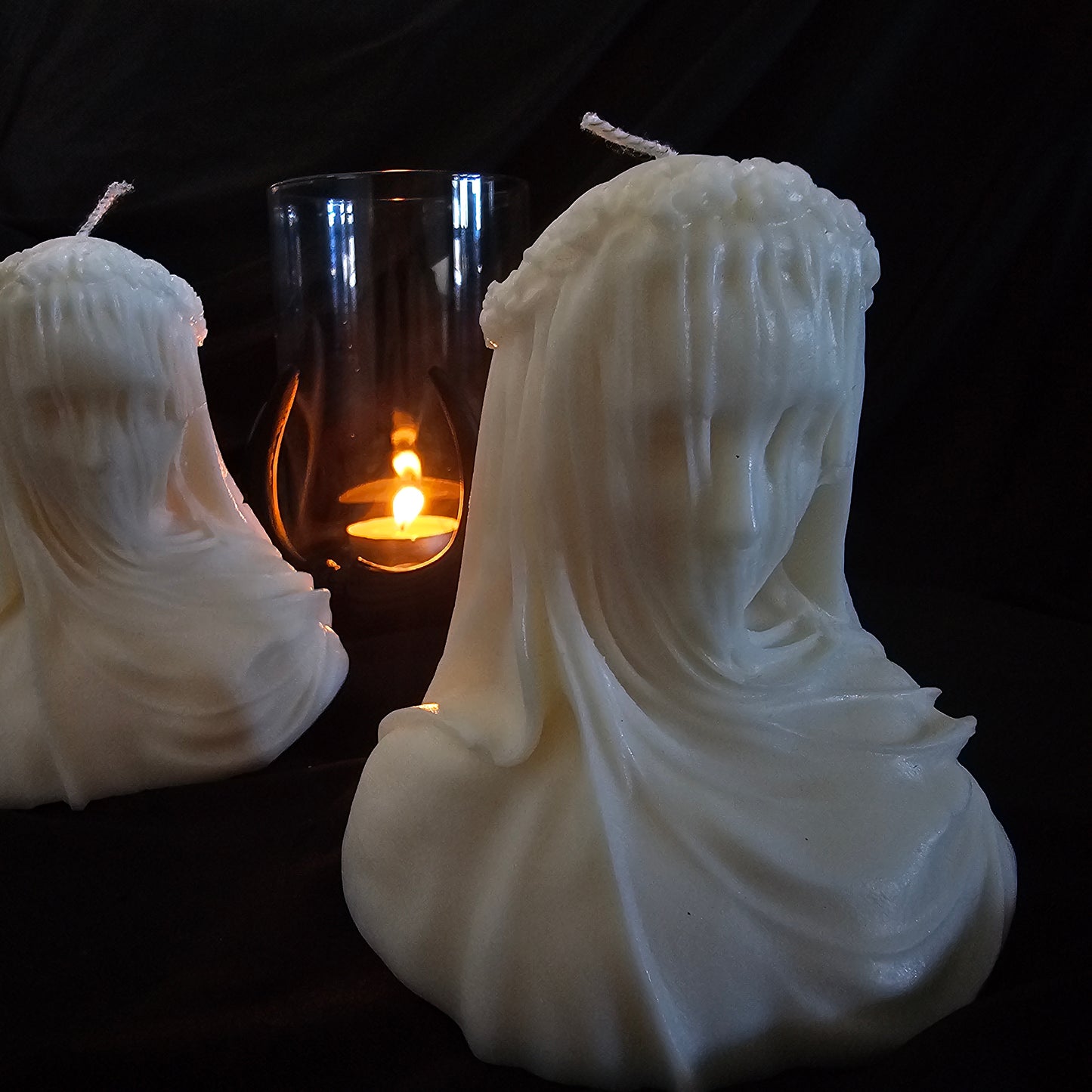 Two off-white candles designed to resemble figures with veils, with a smaller lit candle in the background creating a warm glow.