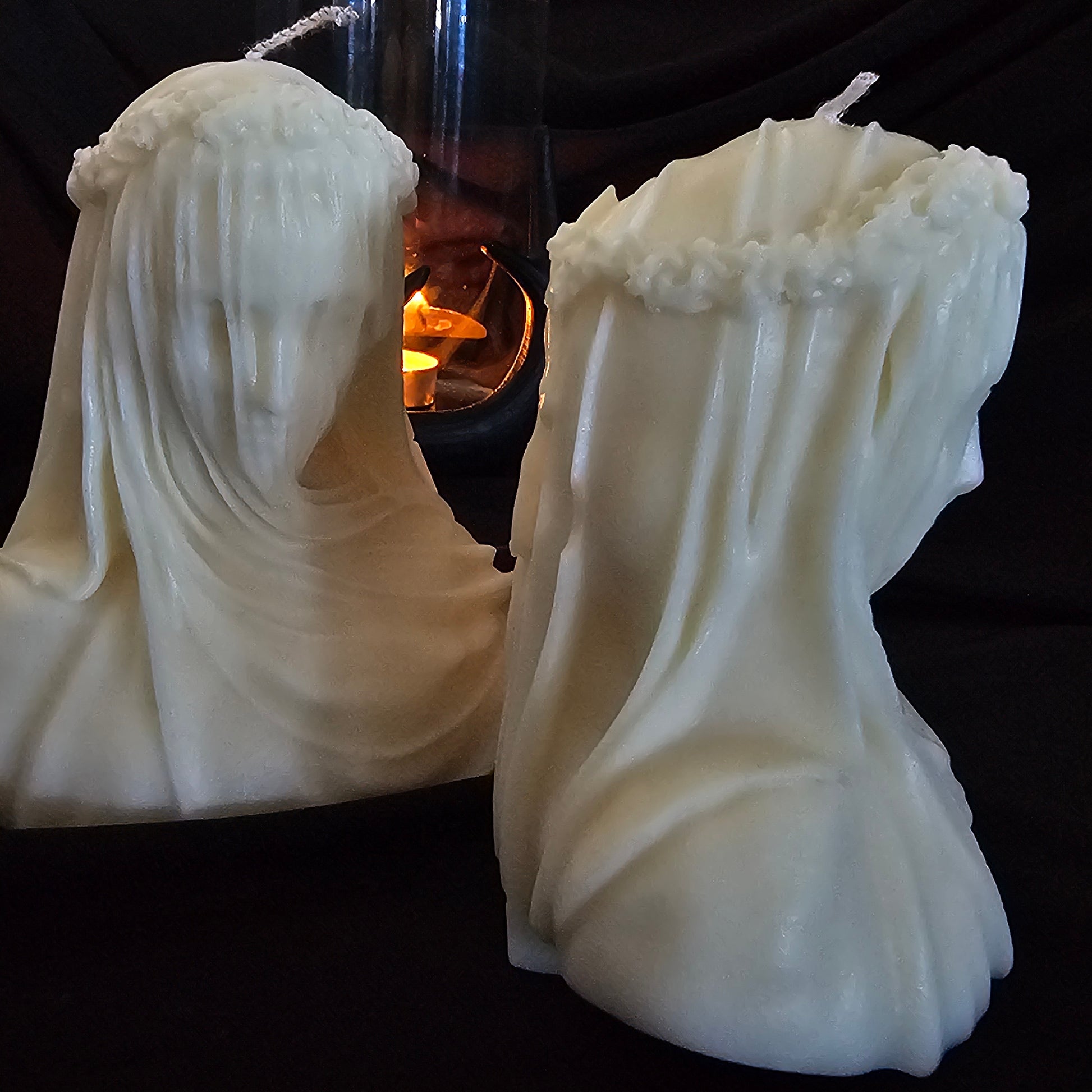 Two off-white candles designed to resemble figures with veils, with a smaller lit candle in the background creating a warm glow.