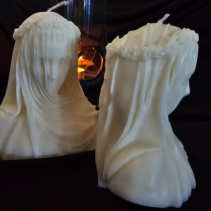 Two off-white candles designed to resemble figures with veils, with a smaller lit candle in the background creating a warm glow.