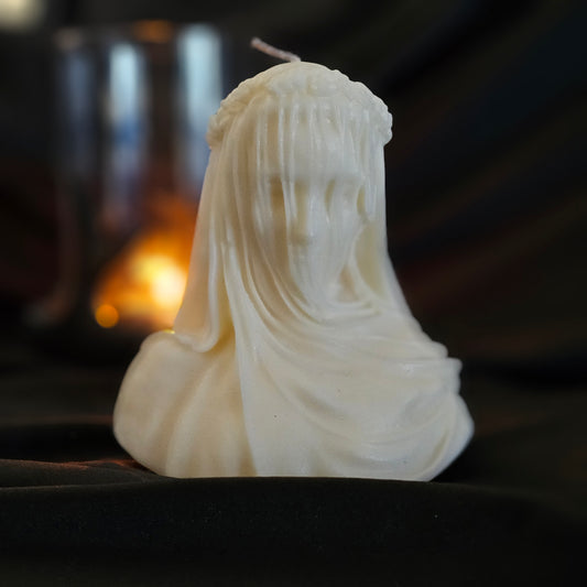 Off-white candle designed to resemble figures with veils, with a smaller lit candle in the background creating a warm glow.