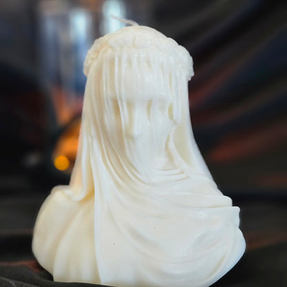 Off-white candle designed to resemble figure with a veil, with a smaller lit candle in the background creating a warm glow.