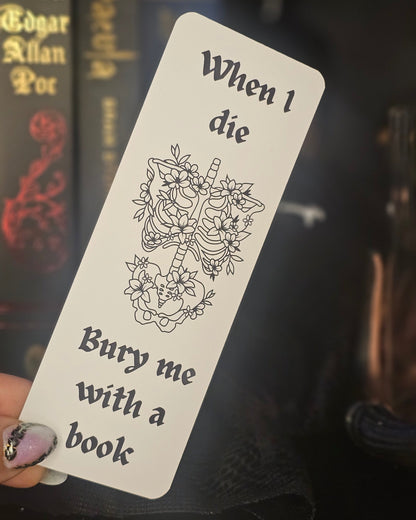 Bookmark with skeleton and floral design, text 'When I die, bury me with a book', held against a blurred bookshelf background.