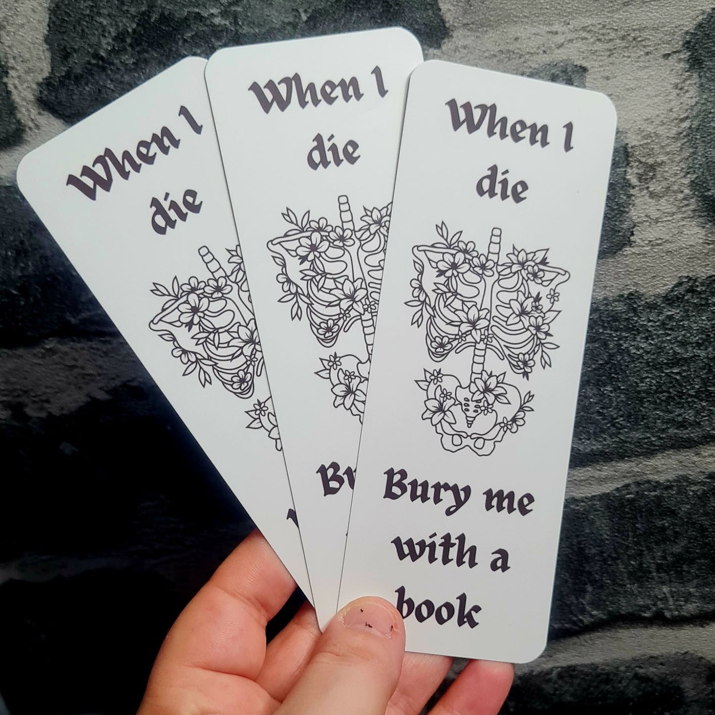 Bookmark with skeleton and floral design, text 'When I die, bury me with a book', held against stonework 