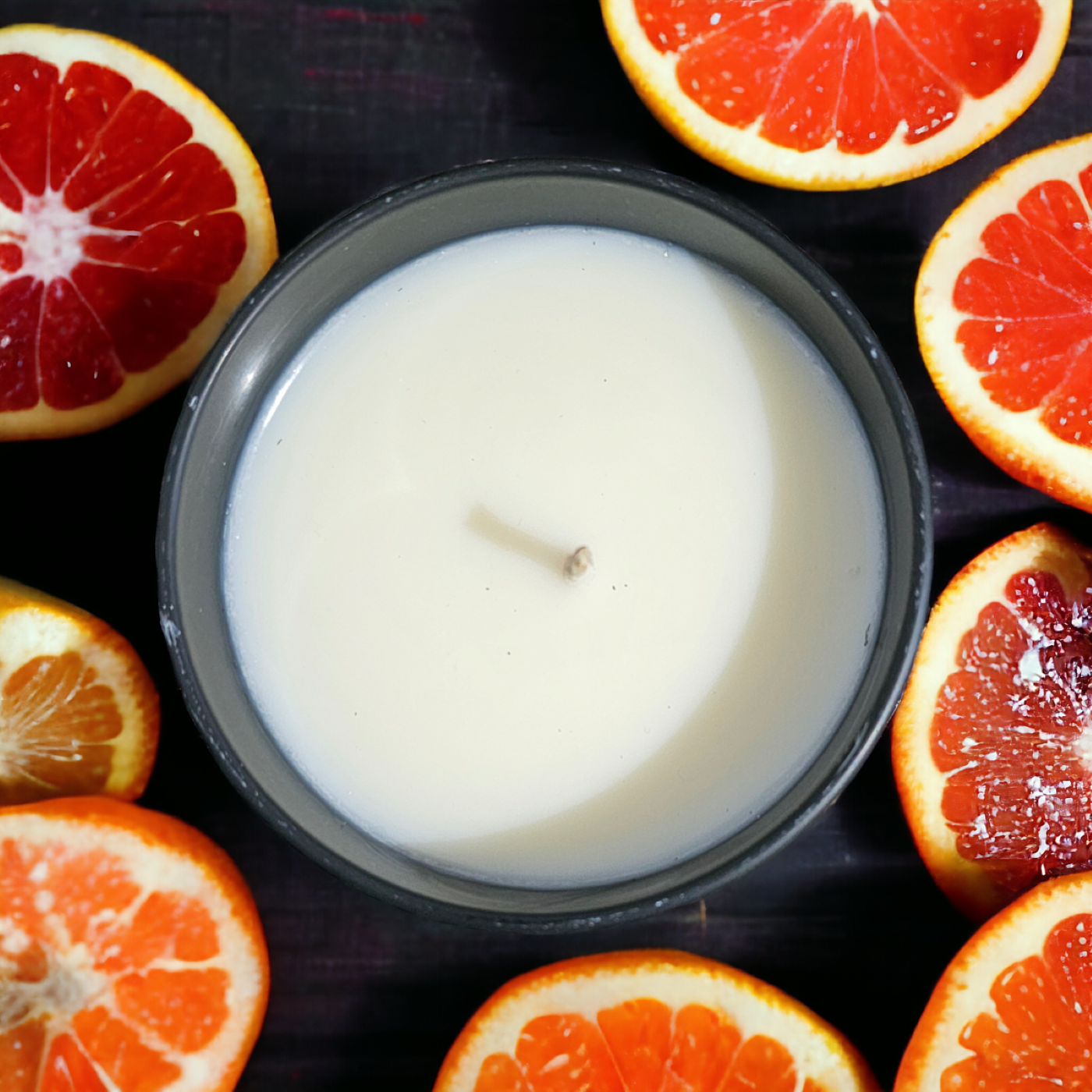A white candle housed in a black glass container, surrounded by sliced blood oranges.