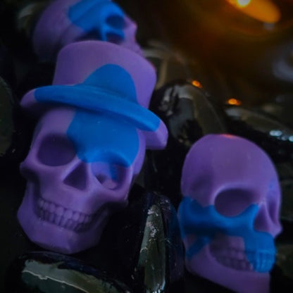 Two purple and blue coloured Skull shaped wax melts on a reflective black surface 