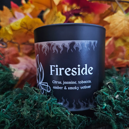 A black glass candle container with a white bamboo lid, labeled 'Fireside' with the scents listed as citrus, jasmine, tobacco, amber, and smoky vetiver, surrounded by autumn leaves.