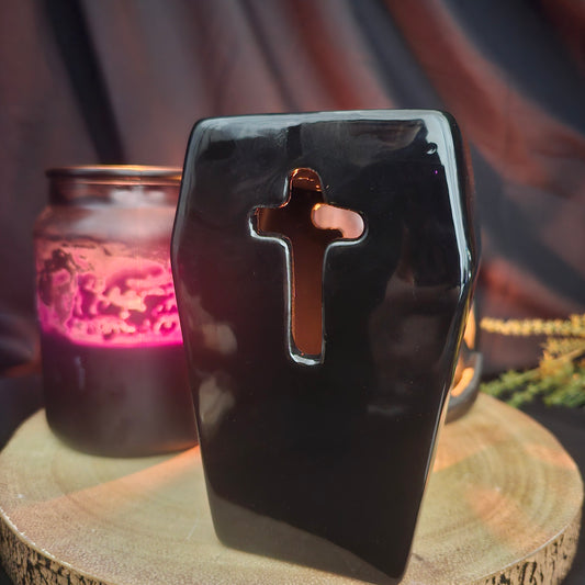 A black ceramic coffin-shaped oil and wax melt burner with a cut out cross design on the front, placed on a wooden surface with a lit candle in the background.