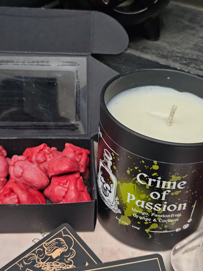 A candle housed in a black jar labelled "Crime of Passion; mango, passionfruit, Orange & Coconut" sitting beside a box of anatomical heart shaped wax melts