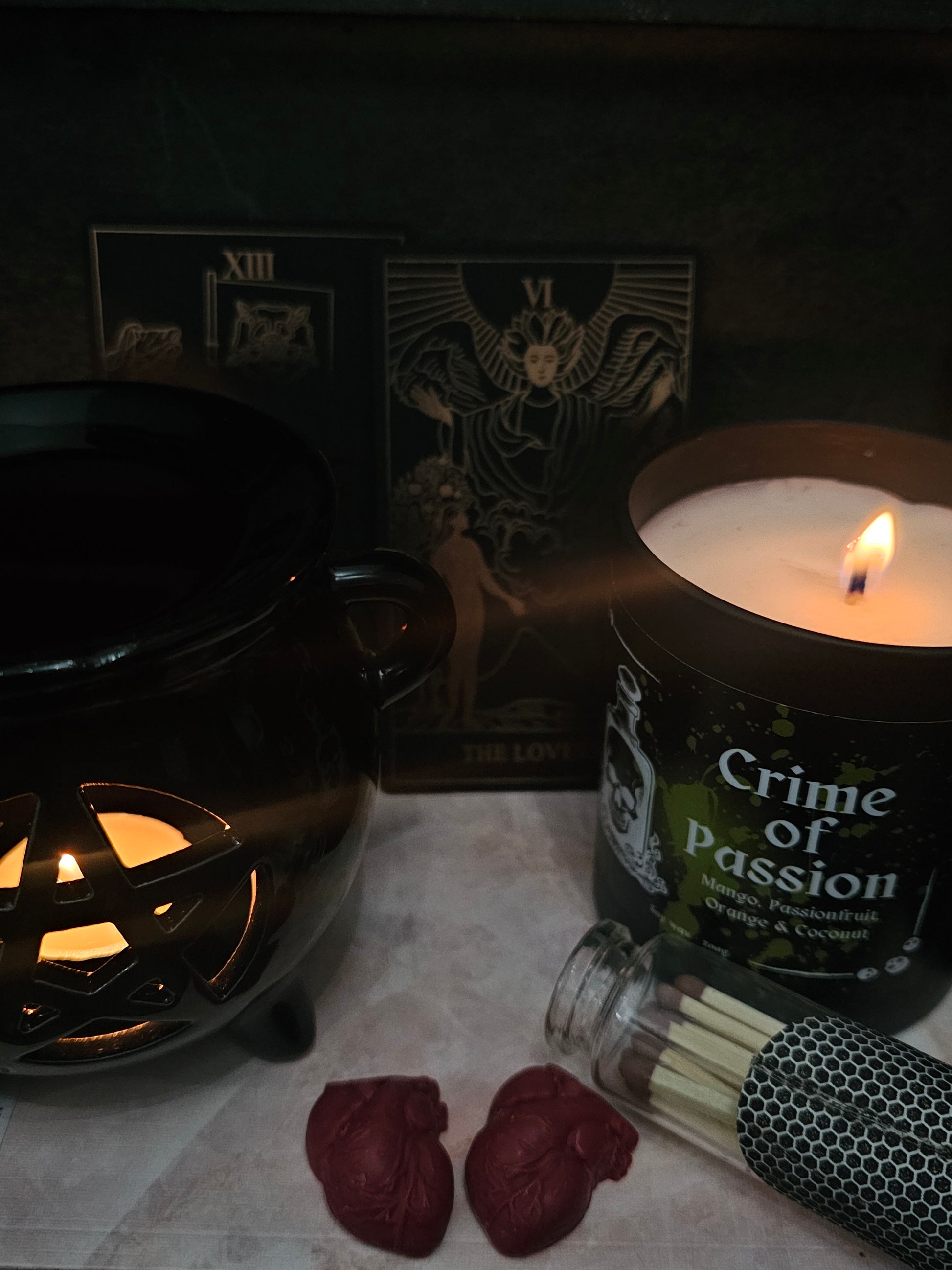 Candle labeled 'Crime of Passion' sitting on a marble surface with a lit wax burner, anitomical heart shaped melts, and "the lovers" tarot card in the background