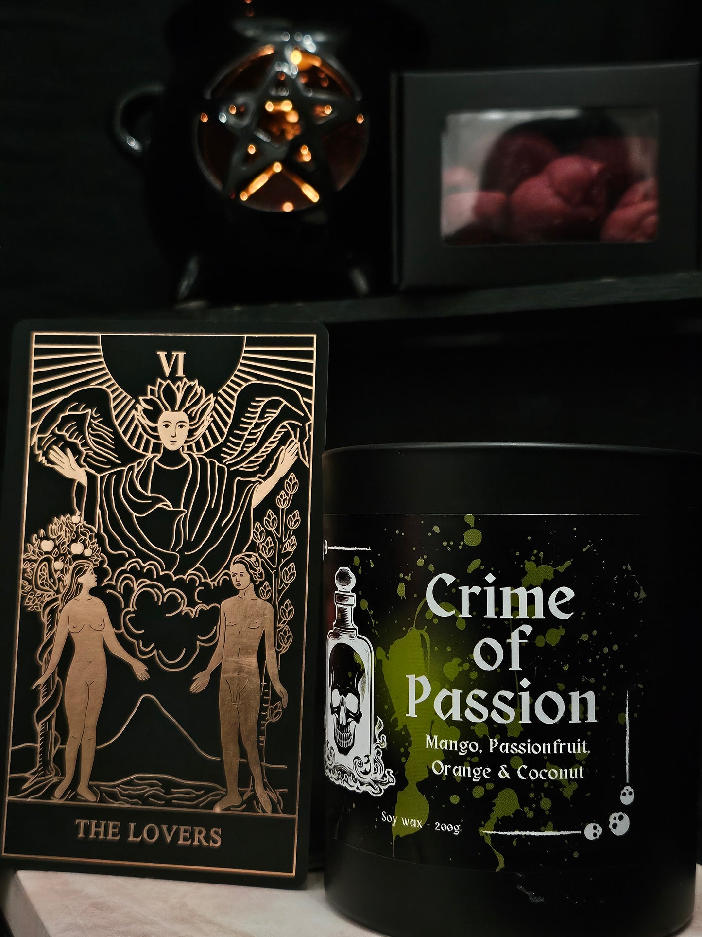 Candle labeled 'Crime of Passion' sitting on a marble surface, in the background a pentagram shaped wax burner and melts, and "the lovers" tarot card 