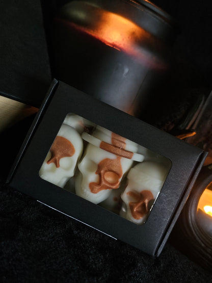 A box containing seven skull-shaped wax melts with a honeycomb pattern on the surface, displayed in a dark setting with candlelight.