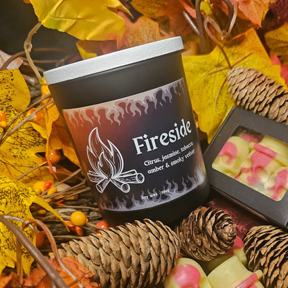 A black glass candle container with a white bamboo lid, labeled 'Fireside' with the scents listed as citrus, jasmine, tobacco, amber, and smoky vetiver, surrounded by autumn leaves.