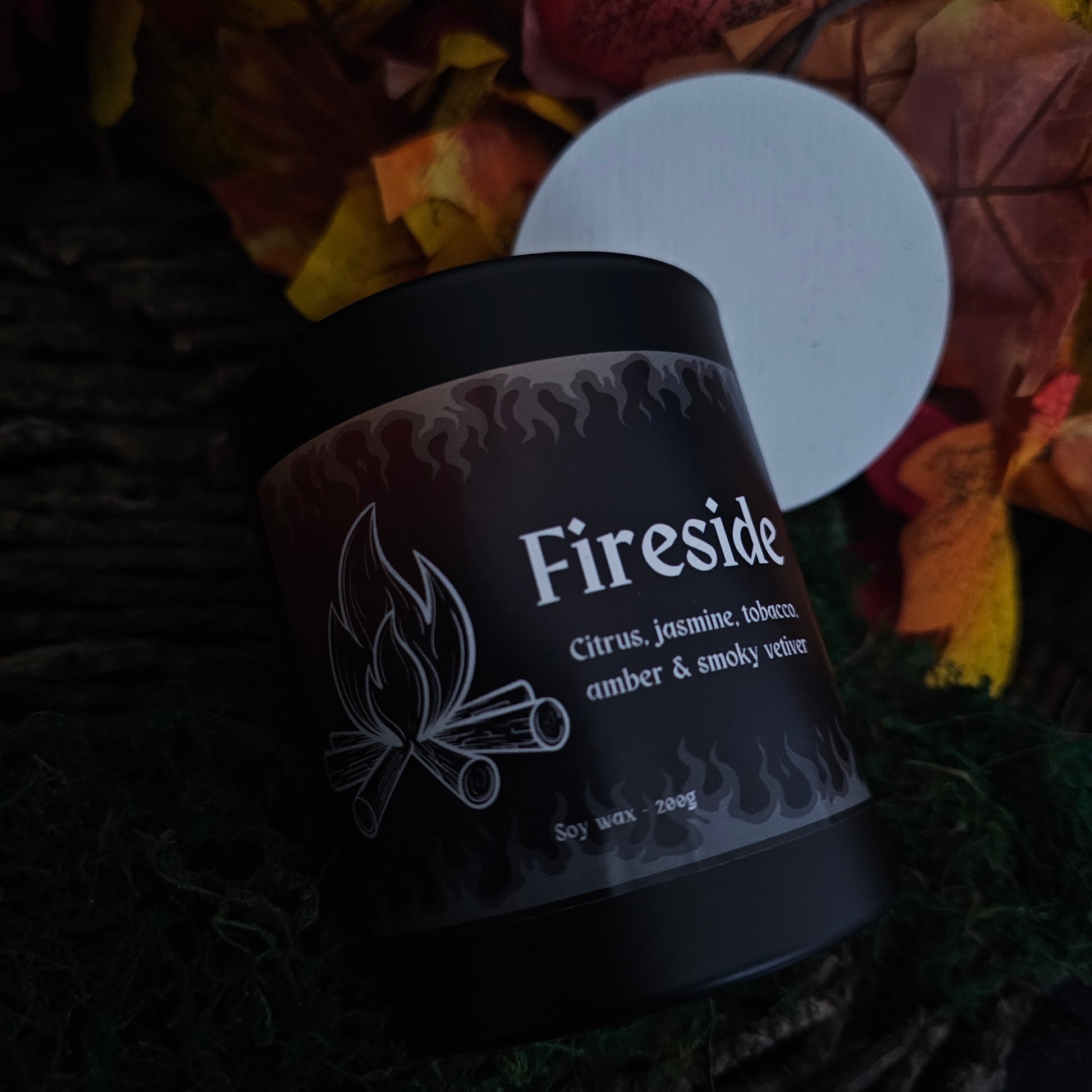 A black glass candle container with a white bamboo lid, labeled 'Fireside' with the scents listed as citrus, jasmine, tobacco, amber, and smoky vetiver, surrounded by autumn leaves.