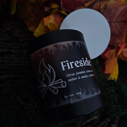 A black glass candle container with a white bamboo lid, labeled 'Fireside' with the scents listed as citrus, jasmine, tobacco, amber, and smoky vetiver, surrounded by autumn leaves.