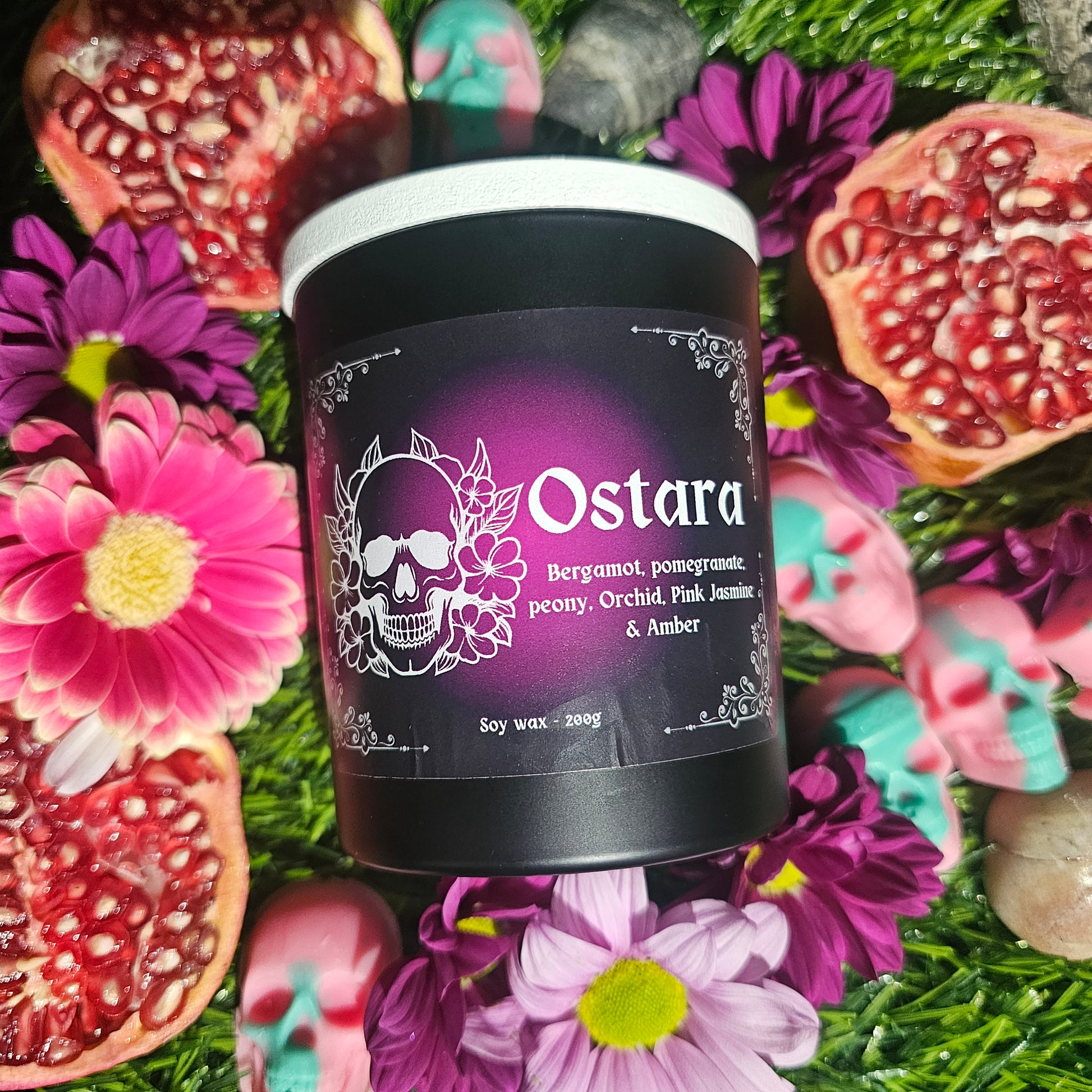 A black glass candle container with a white lid, labeled 'Ostara' with a graphic of a skull and flowers, surrounded by pomegranates and flowers on a green background.