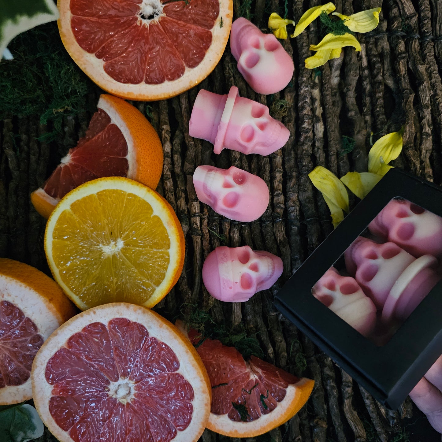Various skull shaped wax melts of pinkish hue lying on twigs surrounded by moss and blood orange slices 