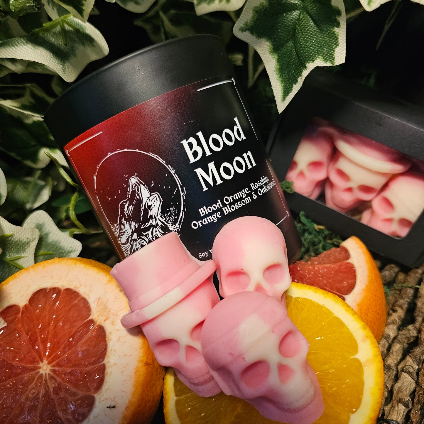 Dark Container Candle labelled "Blood moon" and Skull shaped wax melts sitting on a dark background surrounded by blood orange and ivy leaves 