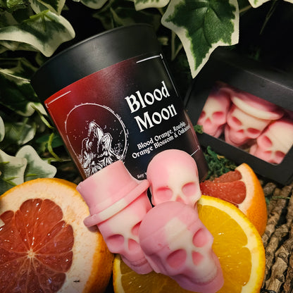 Dark Container Candle labelled "Blood moon" and Skull shaped wax melts sitting on a dark background surrounded by blood orange and ivy leaves 