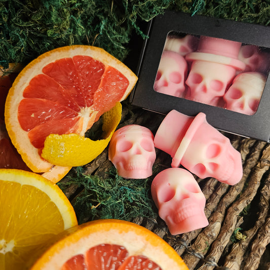 A set of skull-shaped wax melts with a pink hue, displayed alongside sliced blood orange, with a dark, textured background.