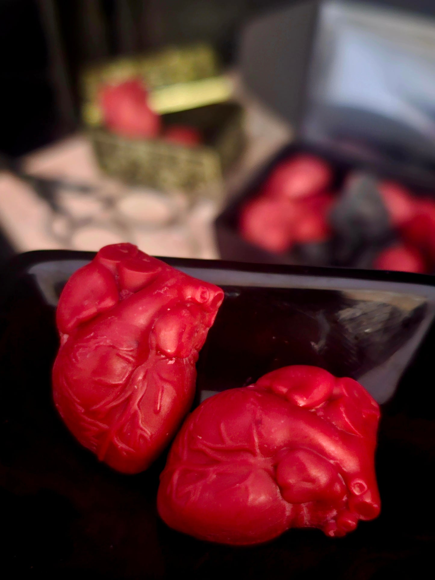 Up close photograph of two anitomical heart shaped wax melts on a black surface with a box of the product blurred background 