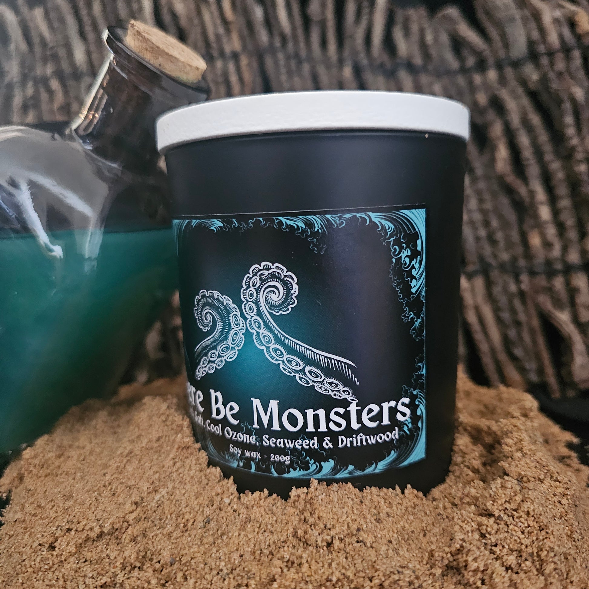 A candle in a black container with "here be monsters" written on a blue and black label. Sitting with a bottle on a sandy surface 