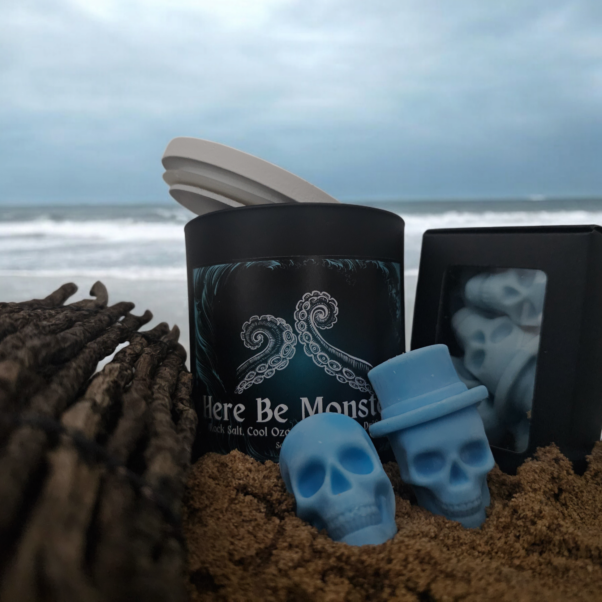 A black candle with a label that reads "Here be monsters" placed on a sandy surface with a bottle and blue skull shaped wax melts with the ocean in the background.