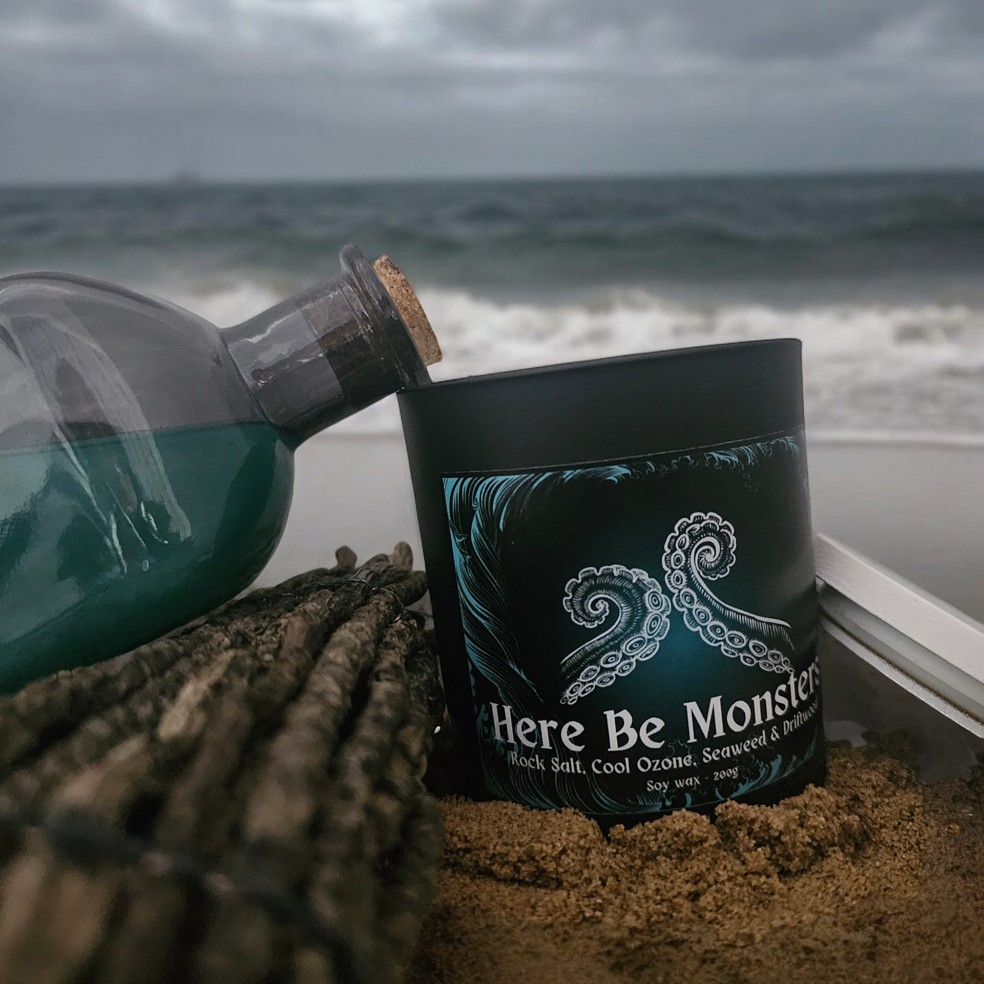 A black candle with a label that reads "Here be monsters" placed on a sandy surface with a bottle and the ocean in the background.