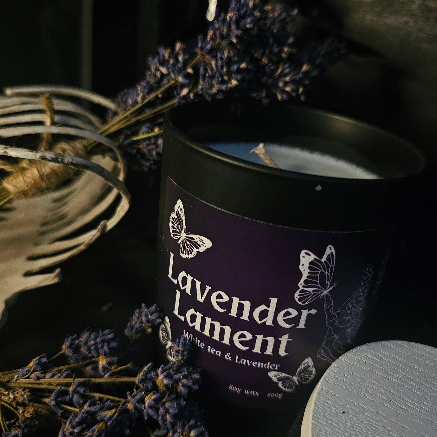 A black glass candle container with a white bamboo lid, labeled 'Lavender Lament', surrounded by dried lavender flowers.