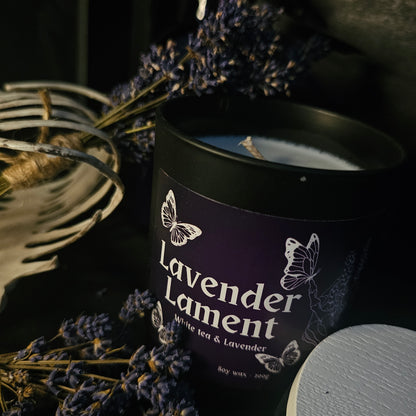 A black glass candle container with a white bamboo lid, labeled 'Lavender Lament', surrounded by dried lavender flowers.