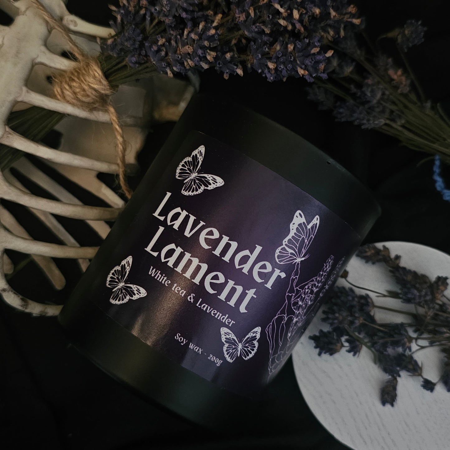 A candle in a black container labeled "Lavender lament; White tea and lavender" on a dark background with lavender buds and a some bones 