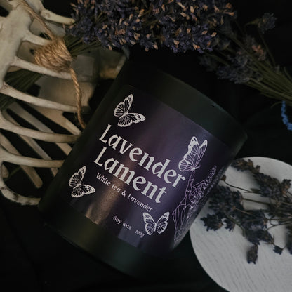 A candle in a black container labeled "Lavender lament; White tea and lavender" on a dark background with lavender buds and a some bones 