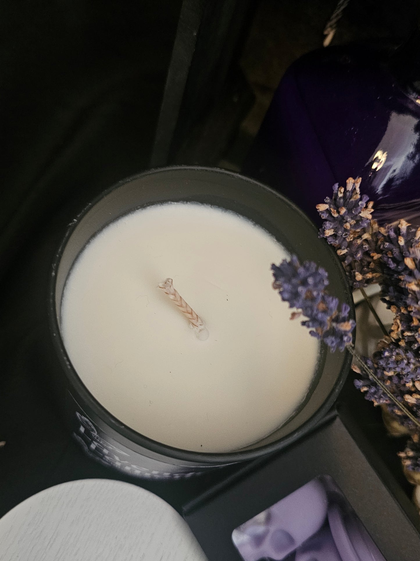 a candle in a black container on a dark background with lavender buds and a purple bottle 