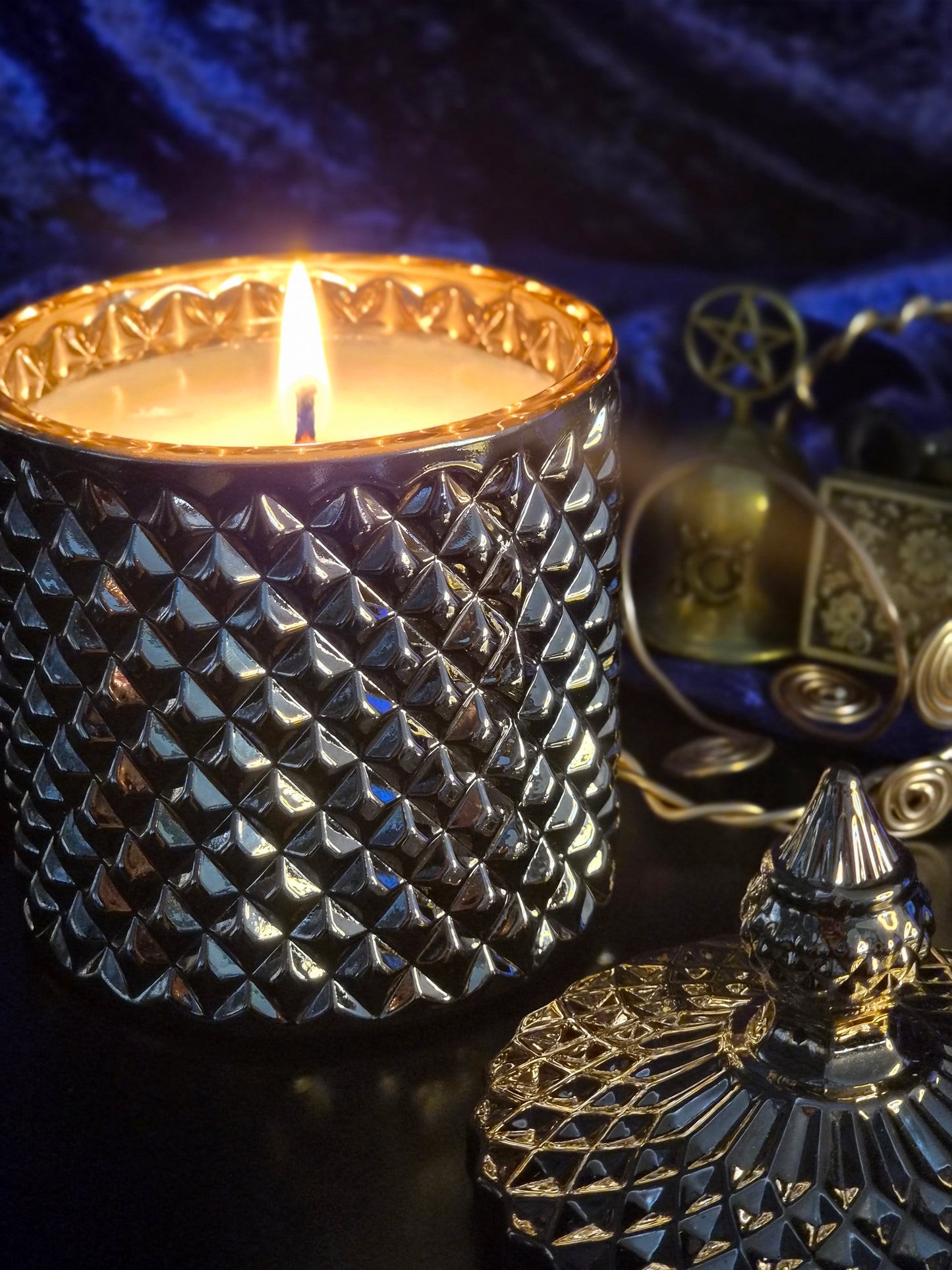 A lit candle in a textured, diamond-patterned holder with a glowing flame, set against a dark blue velvet background with occult-themed decorative props.